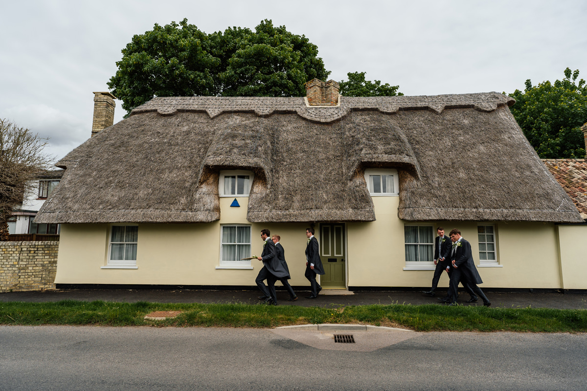 groomsmen walk to the church and pass a thatched cottage in Cambridgeshire village