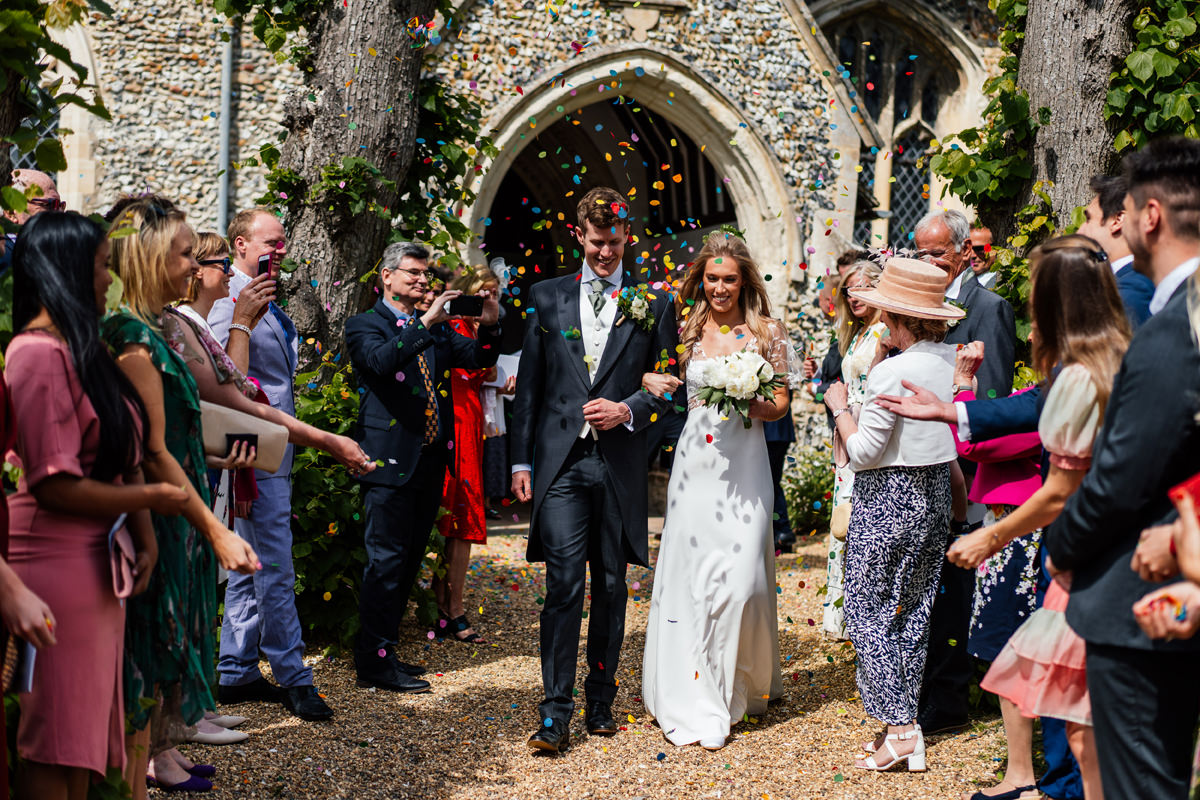 bride and groom are showered with confetti as the walk down the church pathway in full bright sunshine